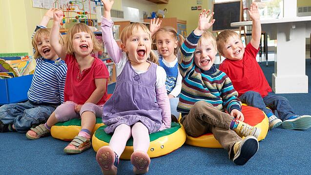Gruppe Kinder sitzt auf Sitzkissen und hebt lachend die Hand.