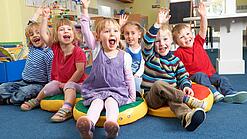 Gruppe Kinder sitzt auf Sitzkissen und hebt lachend die Hand.