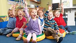 Gruppe Kinder sitzt auf Sitzkissen und hebt lachend die Hand.