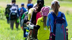 Grundschulkinder wandern mit Rucksäcken über eine grüne Wiese.