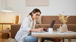 Junge Frau sitzt auf dem Boden vor einem Laptop und notiert etwas.