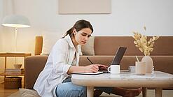 Junge Frau sitzt auf dem Boden vor einem Laptop und notiert etwas.