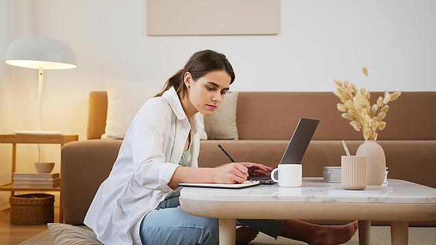 Junge Frau sitzt auf dem Boden vor einem Laptop und notiert etwas.