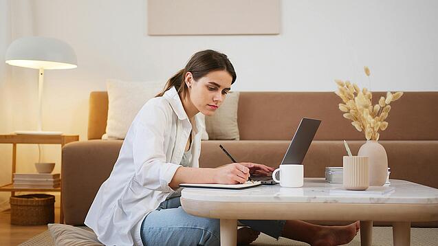 Junge Frau sitzt auf dem Boden vor einem Laptop und notiert etwas.