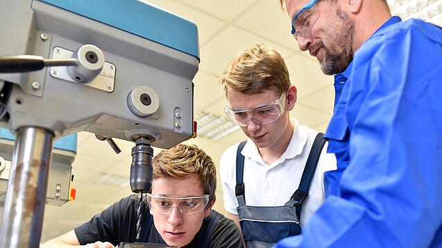 Zwei Jugendliche und ein Erwachsener stehen in Arbeitskleidung an einer technischen Maschine.