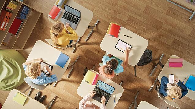 Kinder sitzen in einem Klassenzimmer und arbeiten mit verschiedenen digitalen Geräten.