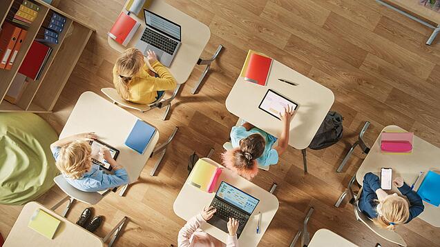 Kinder sitzen in einem Klassenzimmer und arbeiten mit verschiedenen digitalen Geräten.
