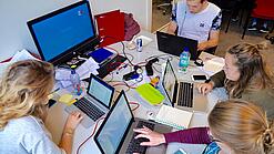 Vier junge Personen sitzen an einem Tisch. Auf dem Tisch stehen Laptops.