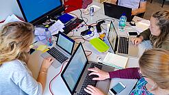 Vier junge Personen sitzen an einem Tisch. Auf dem Tisch stehen Laptops.