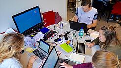 Vier junge Personen sitzen an einem Tisch. Auf dem Tisch stehen Laptops.