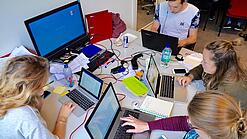 Vier junge Personen sitzen an einem Tisch. Auf dem Tisch stehen Laptops.