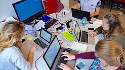 Vier junge Personen sitzen an einem Tisch. Auf dem Tisch stehen Laptops.