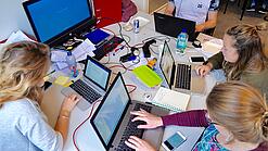 Vier junge Personen sitzen an einem Tisch. Auf dem Tisch stehen Laptops.