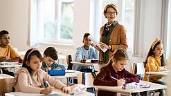 Lehrerin steht in einer Klasse zwischen Schülern, die an Tischen sitzen.