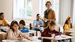 Lehrerin steht in einer Klasse zwischen Schülern, die an Tischen sitzen.