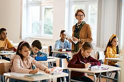 Lehrerin steht in einer Klasse zwischen Schülern, die an Tischen sitzen.