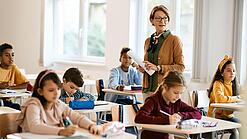 Lehrerin steht in einer Klasse zwischen Schülern, die an Tischen sitzen.