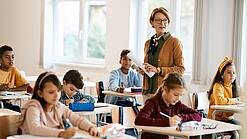 Lehrerin steht in einer Klasse zwischen Schülern, die an Tischen sitzen.