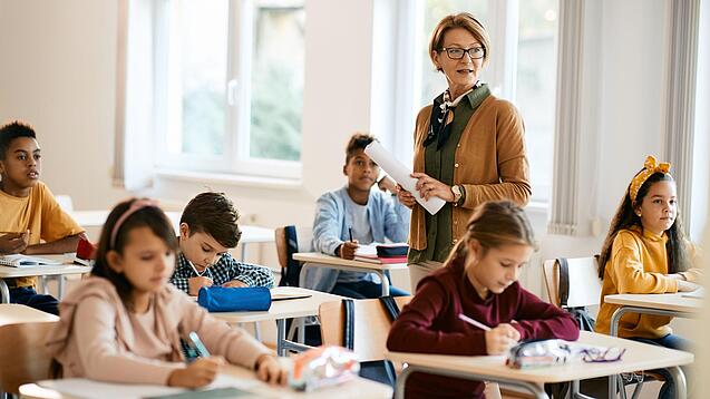 Lehrerin steht in einer Klasse zwischen Schülern, die an Tischen sitzen.