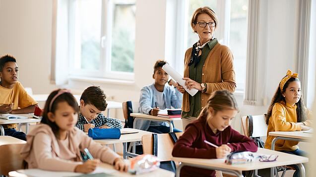 Lehrerin steht in einer Klasse zwischen Schülern, die an Tischen sitzen.