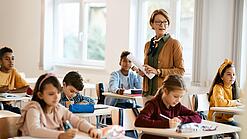Lehrerin steht in einer Klasse zwischen Schülern, die an Tischen sitzen.