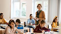 Lehrerin steht in einer Klasse zwischen Schülern, die an Tischen sitzen.