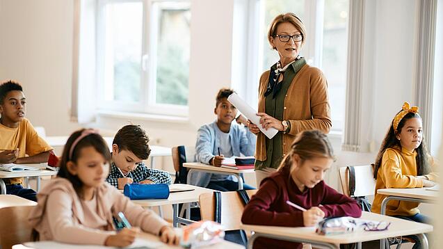 Lehrerin steht in einer Klasse zwischen Schülern, die an Tischen sitzen.