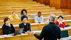 Eine Gruppe studierende in einem Hörsaal. Der Lehrer gestikuliert in Richtung der Gruppe.