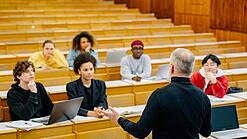 Eine Gruppe studierende in einem Hörsaal. Der Lehrer gestikuliert in Richtung der Gruppe.