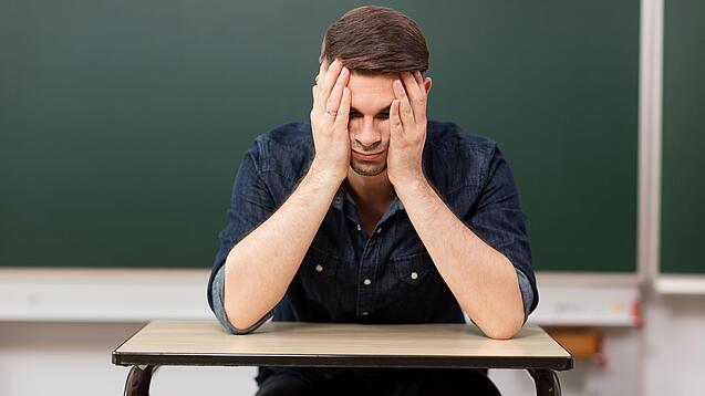 Ein junger Mann sitzt an einem Tisch in einem Klassenzimmer und stützt seinen Kopf auf seine Hände.