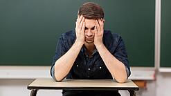 Ein junger Mann sitzt an einem Tisch in einem Klassenzimmer und stützt seinen Kopf auf seine Hände.