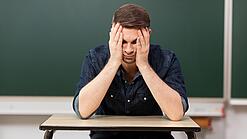 Ein junger Mann sitzt an einem Tisch in einem Klassenzimmer und stützt seinen Kopf auf seine Hände.