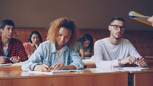 Junge Studierende sitzen in einem Hörsaal bei einer Vorlesung, hören zu und machen Notizen.