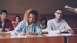 Junge Studierende sitzen in einem Hörsaal bei einer Vorlesung, hören zu und machen Notizen.
