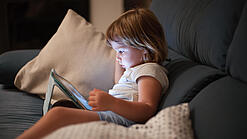 Ein Kleinkind sitzt auf einem Sofa und schaut auf ein Tablet.