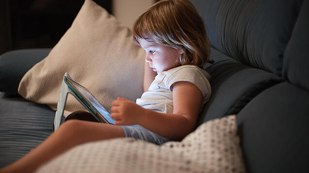 Ein Kleinkind sitzt auf einem Sofa und schaut auf ein Tablet.