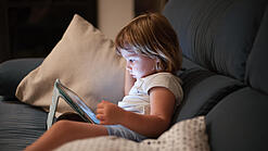 Ein Kleinkind sitzt auf einem Sofa und schaut auf ein Tablet.