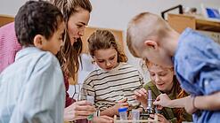 Mehrere Kinder und eine Lehrerin beugen sich über ein Mikroskop.
