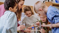 Mehrere Kinder und eine Lehrerin beugen sich über ein Mikroskop.