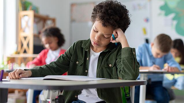 Ein junger Schüler sitzt an einem Schultisch und hat seinen Kopf auf seine Hand gestützt.