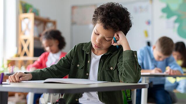 Ein junger Schüler sitzt an einem Schultisch und hat seinen Kopf auf seine Hand gestützt.