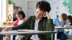 Ein junger Schüler sitzt an einem Schultisch und hat seinen Kopf auf seine Hand gestützt.