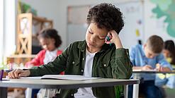 Ein junger Schüler sitzt an einem Schultisch und hat seinen Kopf auf seine Hand gestützt.