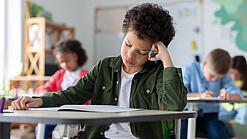Ein junger Schüler sitzt an einem Schultisch und hat seinen Kopf auf seine Hand gestützt.