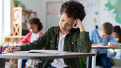 Ein junger Schüler sitzt an einem Schultisch und hat seinen Kopf auf seine Hand gestützt.