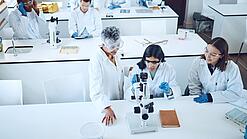 Gruppe Studierender und eine Dozentin in einem Chemiehörsaal vor Mikroskopen