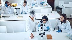 Gruppe Studierender und eine Dozentin in einem Chemiehörsaal vor Mikroskopen