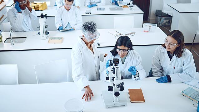 Gruppe Studierender und eine Dozentin in einem Chemiehörsaal vor Mikroskopen