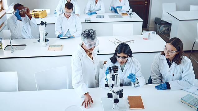 Gruppe Studierender und eine Dozentin in einem Chemiehörsaal vor Mikroskopen