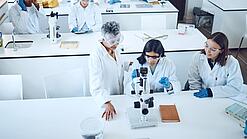 Gruppe Studierender und eine Dozentin in einem Chemiehörsaal vor Mikroskopen